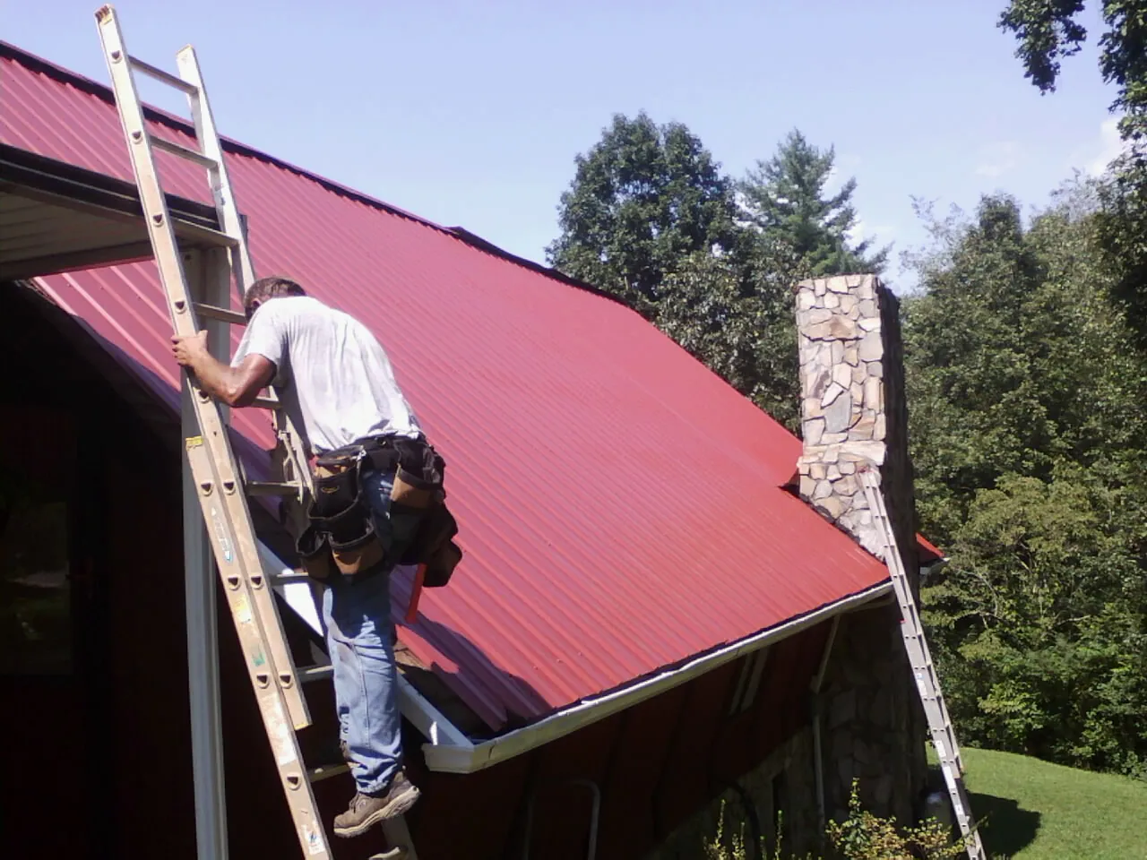 Roofer on a ladder beside a red metal roof and stone chimney — Soffit & Fascia Repair by Roofing Brothers in Loysburg